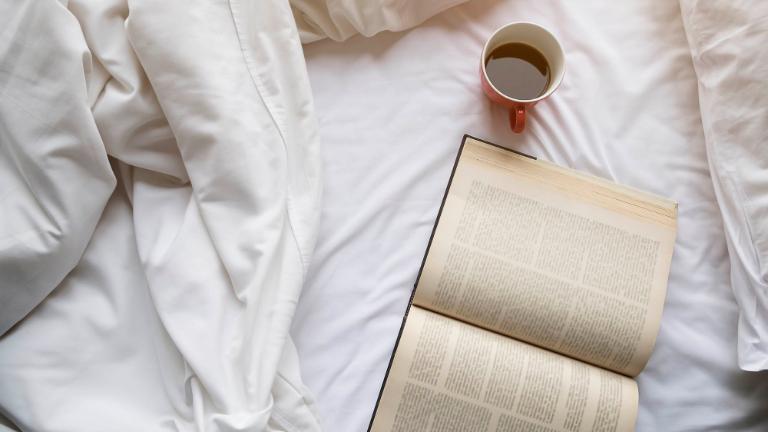 a red cup of coffee, open book, white blanket and white pillow on white bed